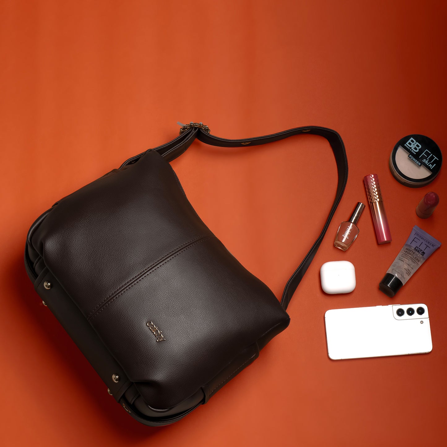 A premium medium-sized brown leather bag lying open on an orange surface, with a phone, earbuds, makeup, and compact powder spilling out—showcasing its spacious design for everyday elegance.