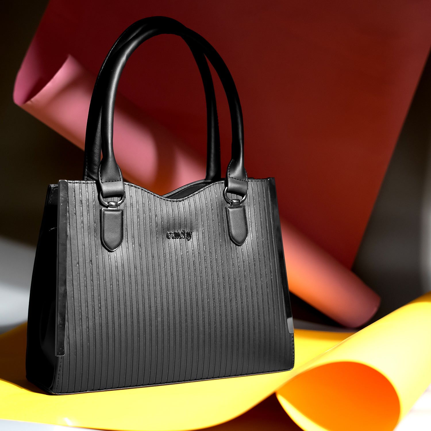 An elegant, lightweight black designer handbag with a top handle, displayed on a yellow surface with a pink and orange paper backdrop—perfect for casual everyday use.